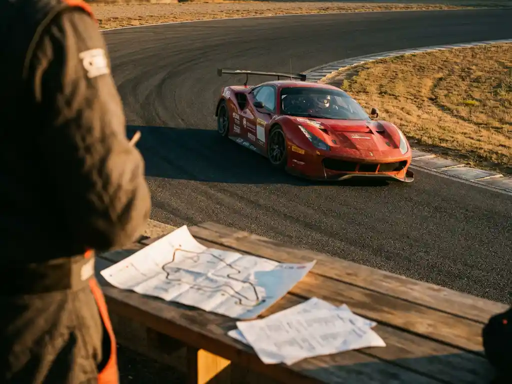 Roter Ferrari in enger Kurve mit sichtbarem Fahrerhelm, Streckenkarte und Notizen auf Pit-Lane-Tisch im Vordergrund.