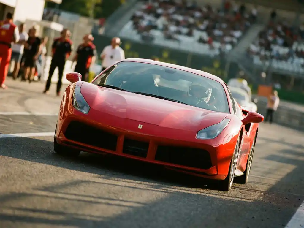 Roter Ferrari-Rennwagen in schneller Kurvenfahrt auf sonnenbeschienem europäischen Rundkurs, Fahrer im Cockpit sichtbar.