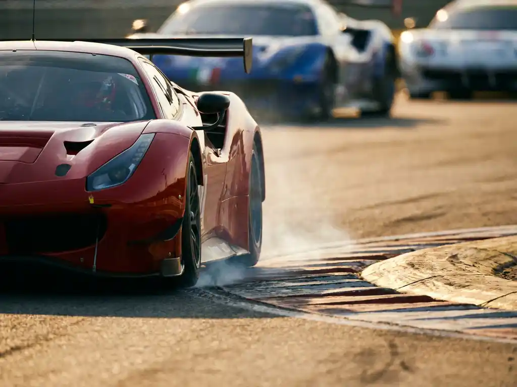 Roter Ferrari-Rennwagen beim Anbremsen einer engen Kurve auf sonnenbeschienem Rundkurs, Vorderreifen unter Last, Rivalen unscharf im Hintergrund.