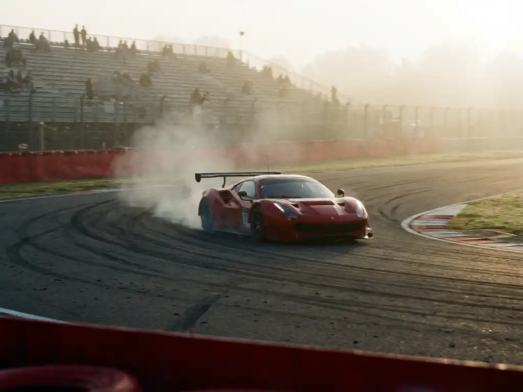 Roter Ferrari-Rennwagen beim Powerslide durch eine Kurve, Reifenrauch und Morgennebel über dem Asphalt, Tribünen im Hintergrund.