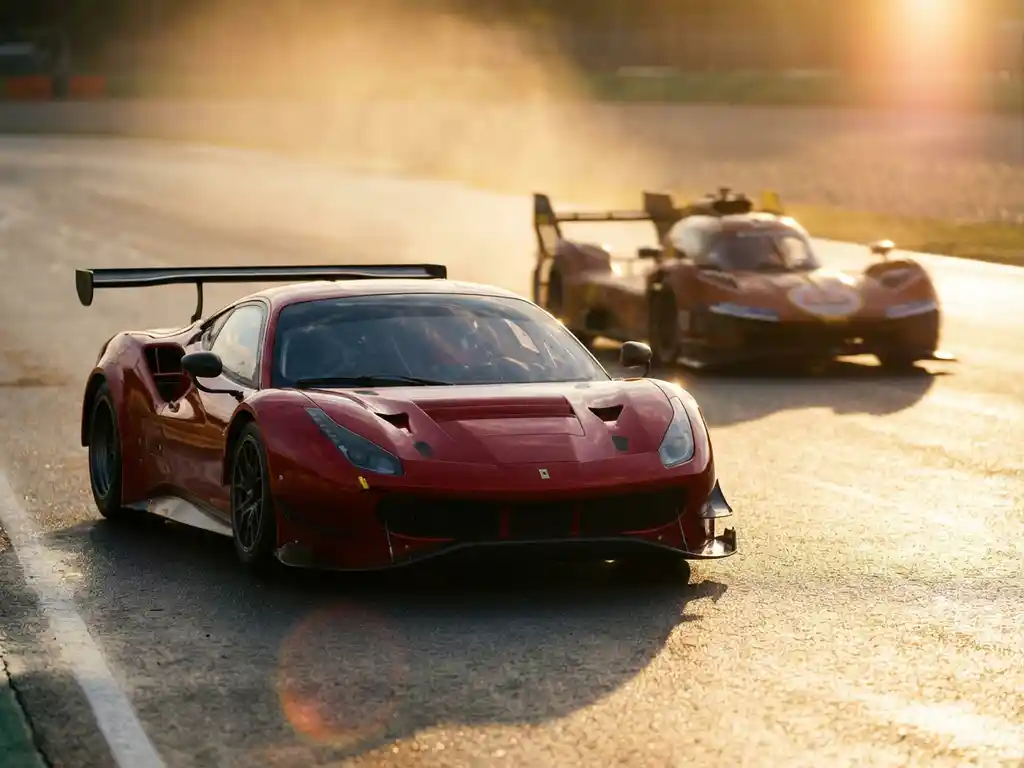 Roter Ferrari-Rennwagen auf nasser Rennstrecke im Sonnenlicht, unscharfer GT-Prototyp im Hintergrund, flache Schärfentiefe.