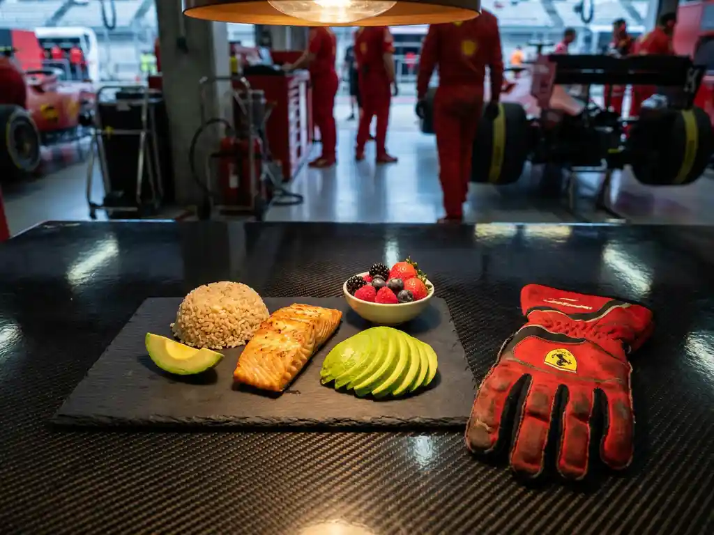Formel-1-Fahrer-Mahlzeit mit gegrilltem Lachs, braunem Reis, Avocado und Beeren neben rotem Ferrari-Rennhandschuh auf Carbonfaser-Oberfläche.