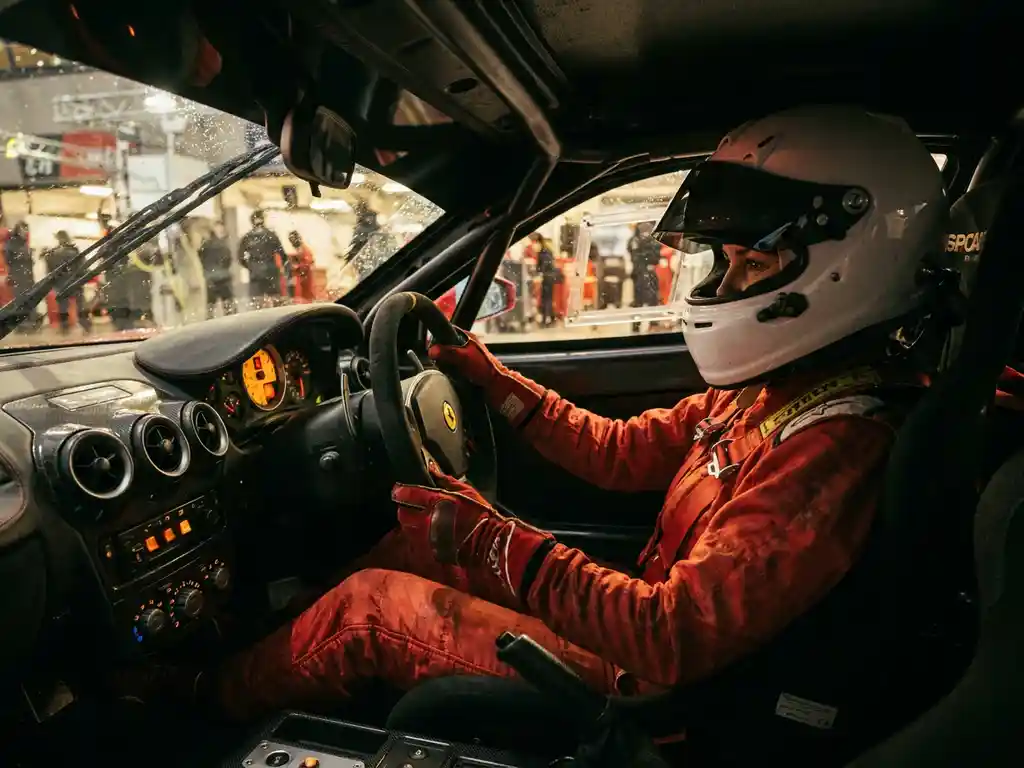 Rennfahrerin in rotem Feuerschutzanzug und weißem Helm am Steuer eines Ferrari GT, Cockpit mit leuchtenden Instrumenten, Boxengasse im Hintergrund.