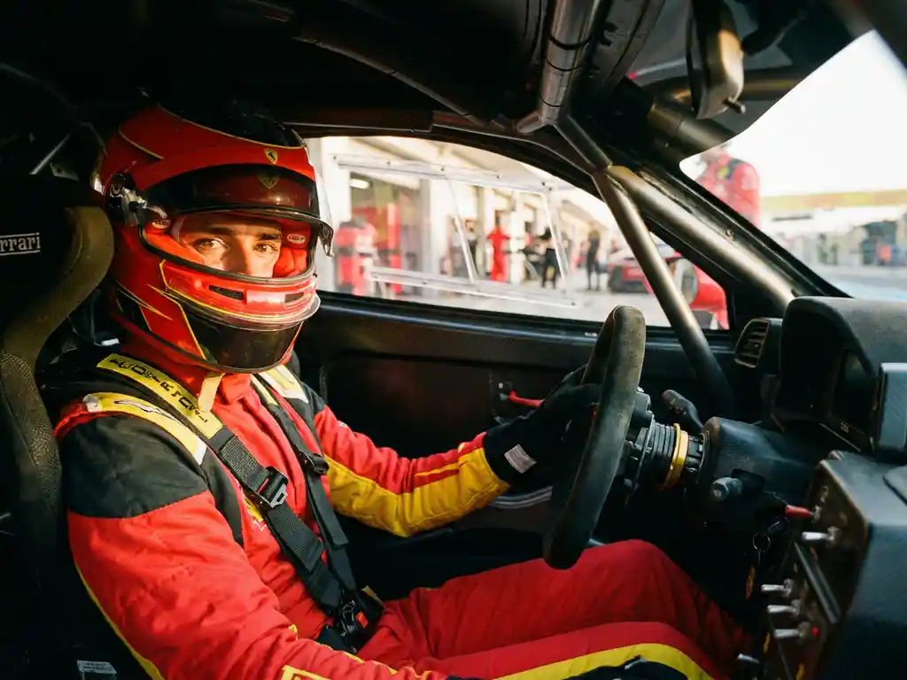 Ferrari-Rennfahrer in rotem Feuerschutzanzug und Helm am Steuer eines Formel-1-Cockpits, Boxengasse im Hintergrund.