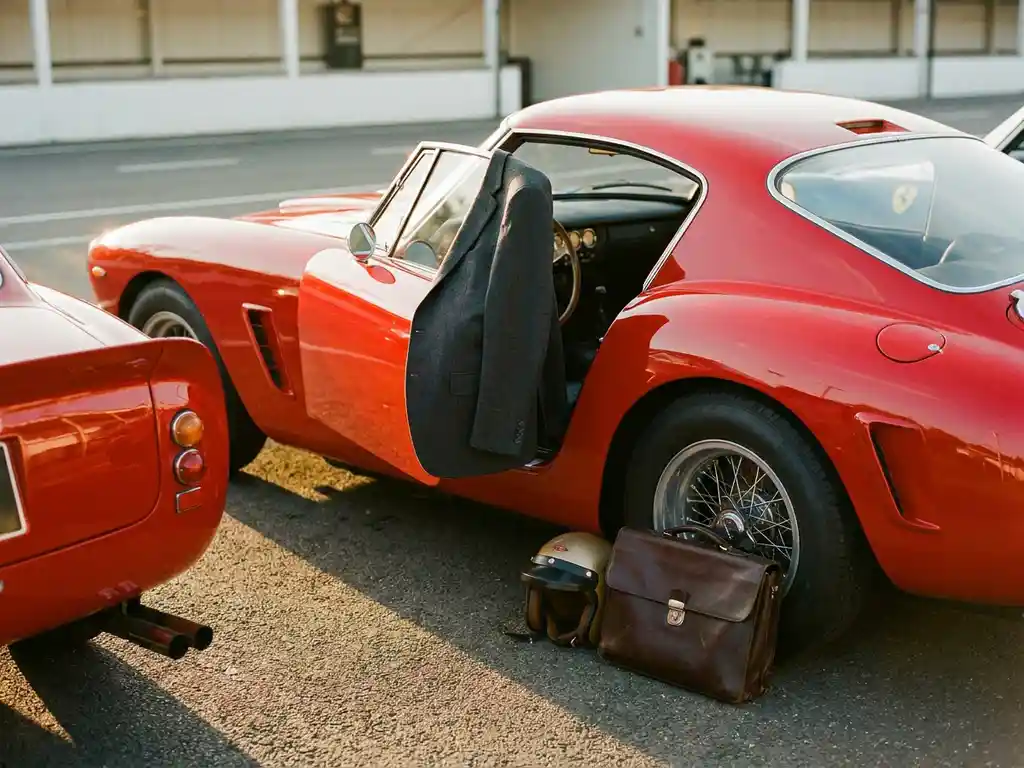 Roter Ferrari in der Boxengasse bei goldenem Abendlicht, mit Businessjacke über der Tür, Lederkoffer und Rennhelm auf dem Asphalt.
